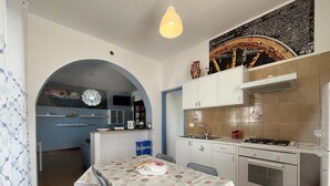 A compact kitchen with a dining area, featuring a central island, a built-in oven, and a gas stove. - PIETRA MARINA APARTMENT (BALESTRATE)