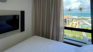 A hotel room with a bed, a flat-screen TV, a window with a view of a pool and beach.