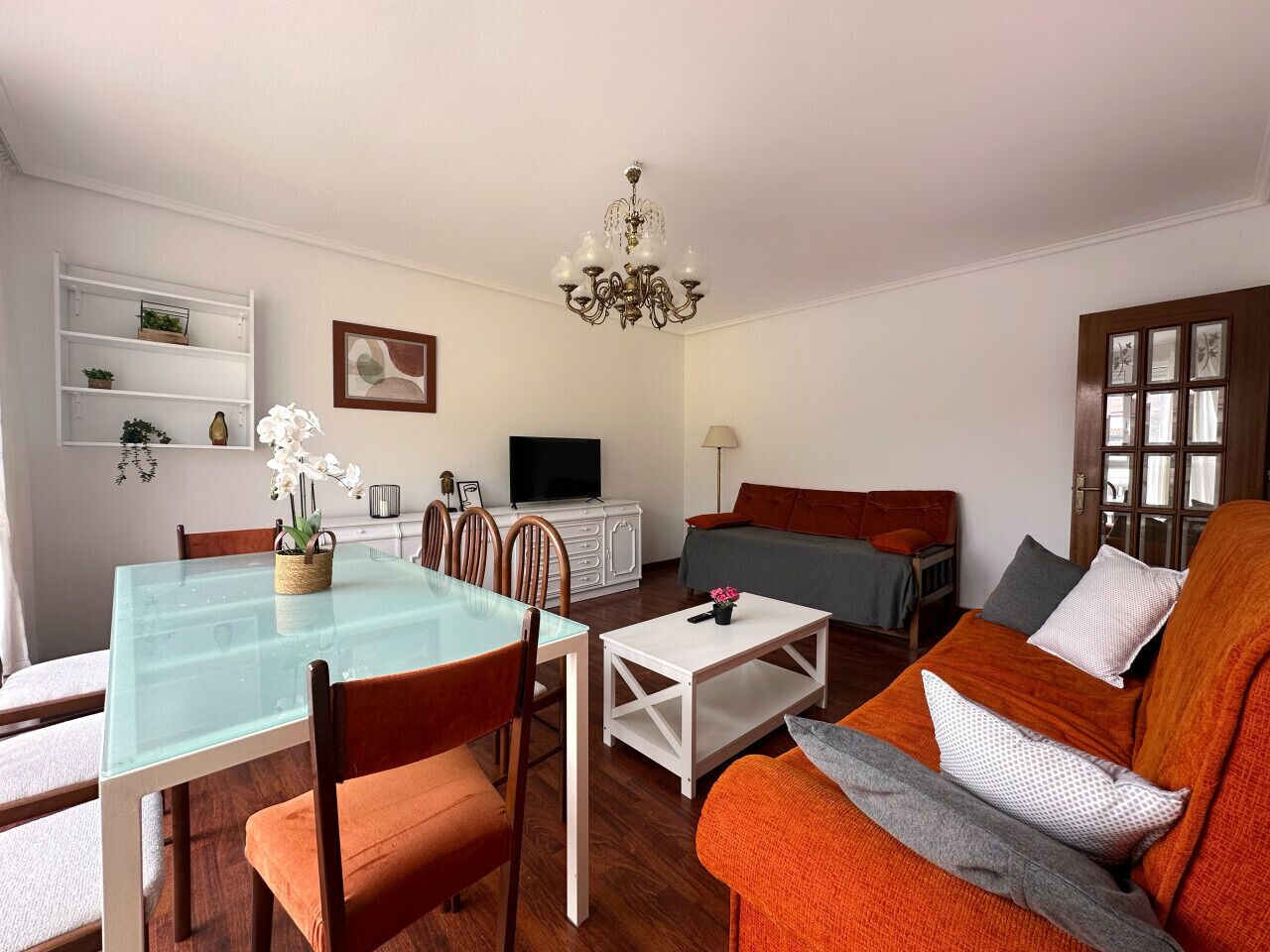 A modern living room with a glass dining table, wooden chairs, a sofa, and a television.