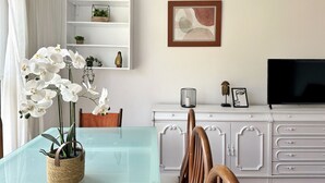 Un comedor con una mesa de vidrio, sillas blancas, un mueble blanco con un televisor y un cuadro abstracto enmarcado en la pared.