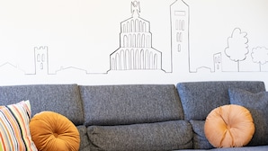 En grå soffa med två runda kuddar, en randig och en enfärgad orange, placerad framför en vägg med en väggmålning som föreställer en stadsbild.