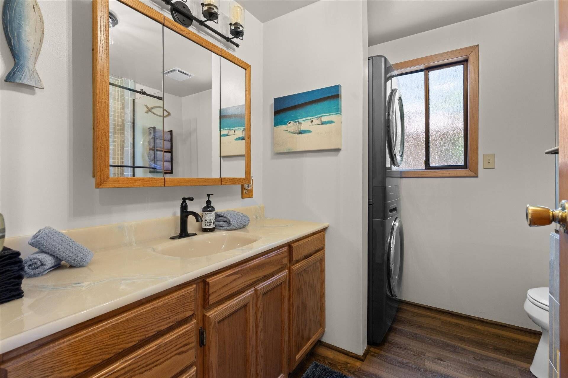 A bathroom with a wooden vanity, a large mirror, a sink with a soap dispenser, and a window with a view of the beach.
