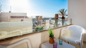 A balcony with a clear view of the cityscape, a white chair, a small table with a plant and two glasses, and a palm tree.