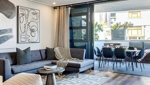 A modern living room with a grey sofa, patterned cushions, a round coffee table, and a large abstract painting. A sliding glass door leads to an outdoor dining area with a table and chairs. - Modern Central Secure Cape Town 2bed Bantry Suite (Cape Town)