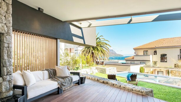 A modern outdoor seating area with a wooden deck, stone walls, and a view of the sea and buildings.