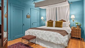 Un dormitorio con una cama con dosel, un banco de madera, una mesita de noche con lámpara y una pequeña mesa con una planta.