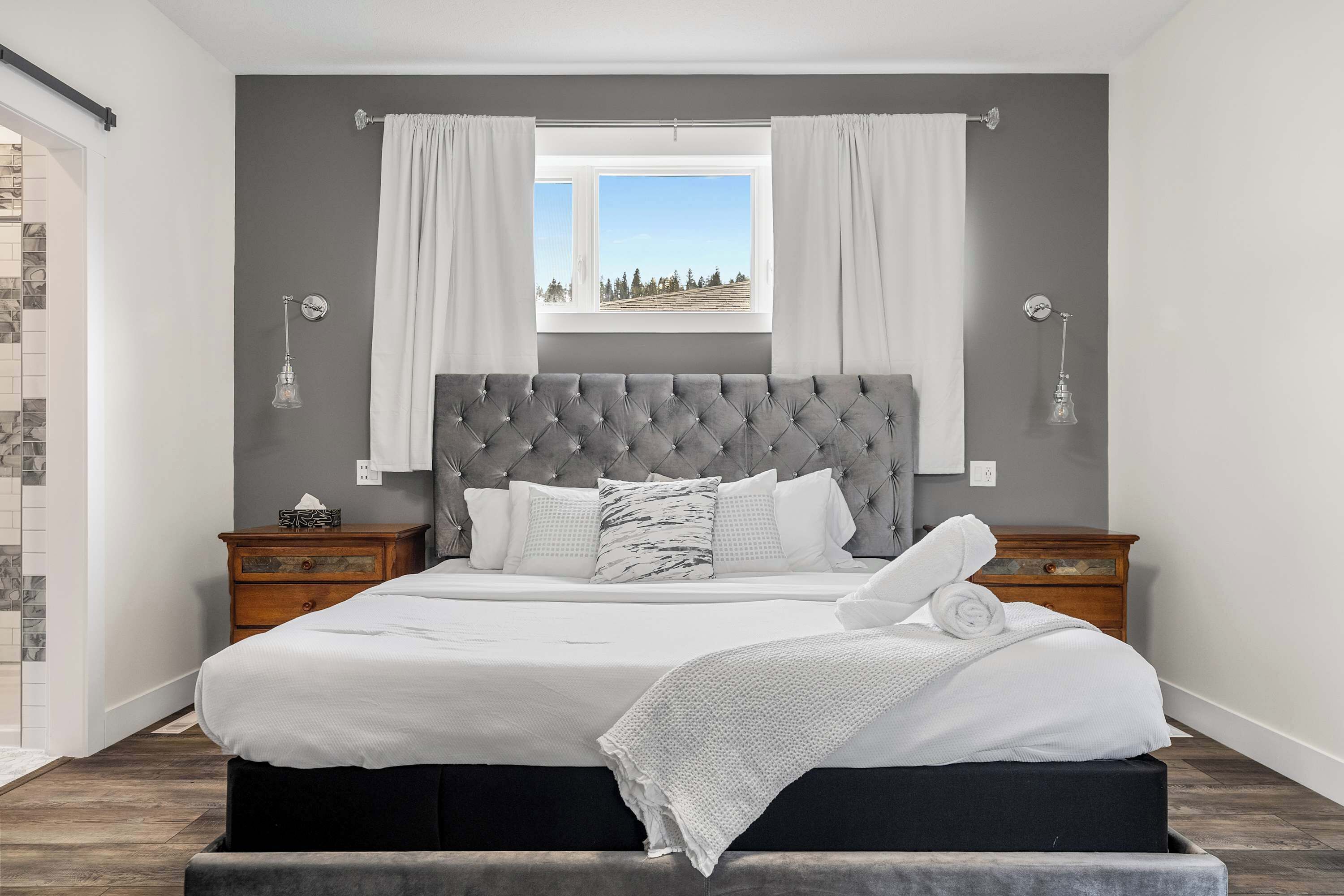 Un dormitorio con una cama grande, cabecera gris, ropa de cama blanca, dos mesitas de noche y un ventanal con cortinas.