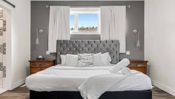 Un dormitorio con una cama grande, cabecera gris, ropa de cama blanca, dos mesitas de noche y un ventanal con cortinas.