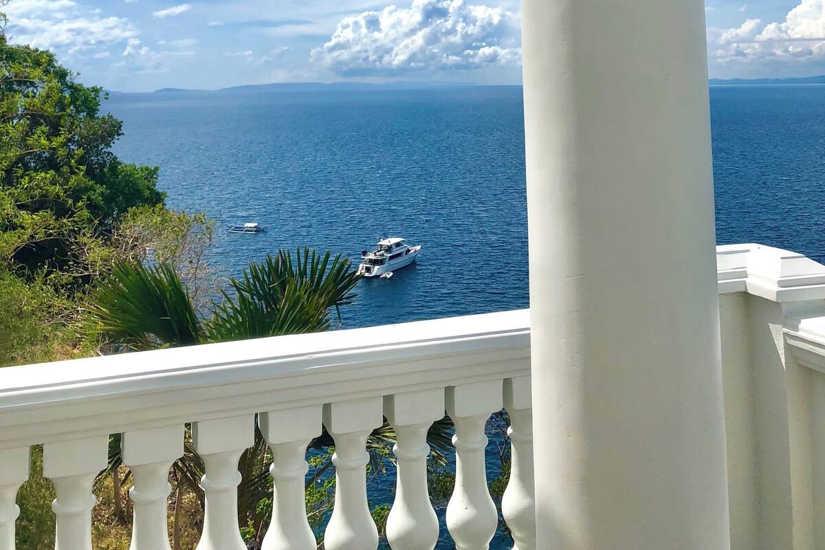 A balcony with a white balustrade overlooking a clear blue sea with boats.