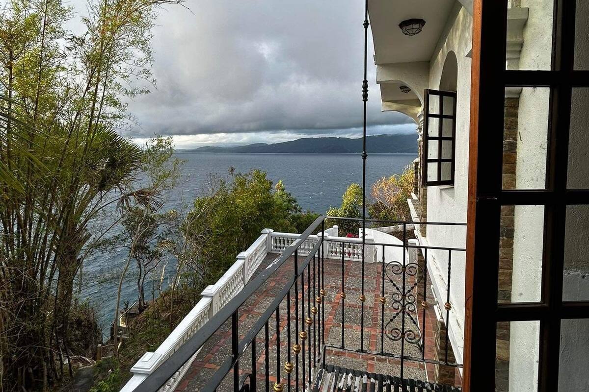 A balcony with a view of the sea and mountains, a railing, and a building with a white facade.