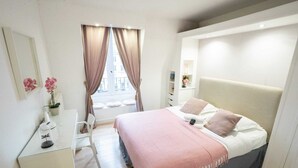 Ein modernes Schlafzimmer mit einem großen Bett, einem Nachttisch, einem Sessel, einem Fenster mit Vorhängen und Blick auf Gebäude.
