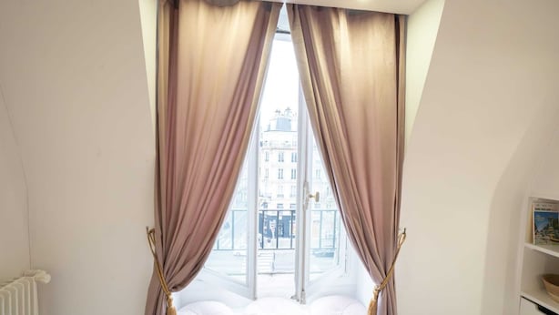 Ein Zimmer mit großem Fenster, Vorhängen, Heizkörper und einem Regal mit einem Buch.