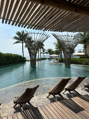 Zona de piscina con sillones de madera, vista despejada al océano y una estructura de palmeras.