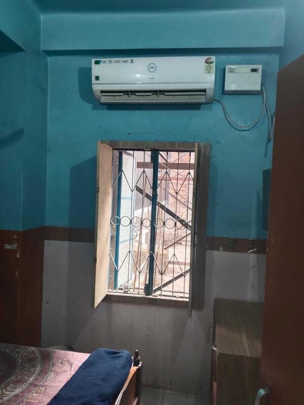 Una habitación pequeña con una cama, un ventanal con rejas, un equipo de aire acondicionado y un enchufe en la pared.