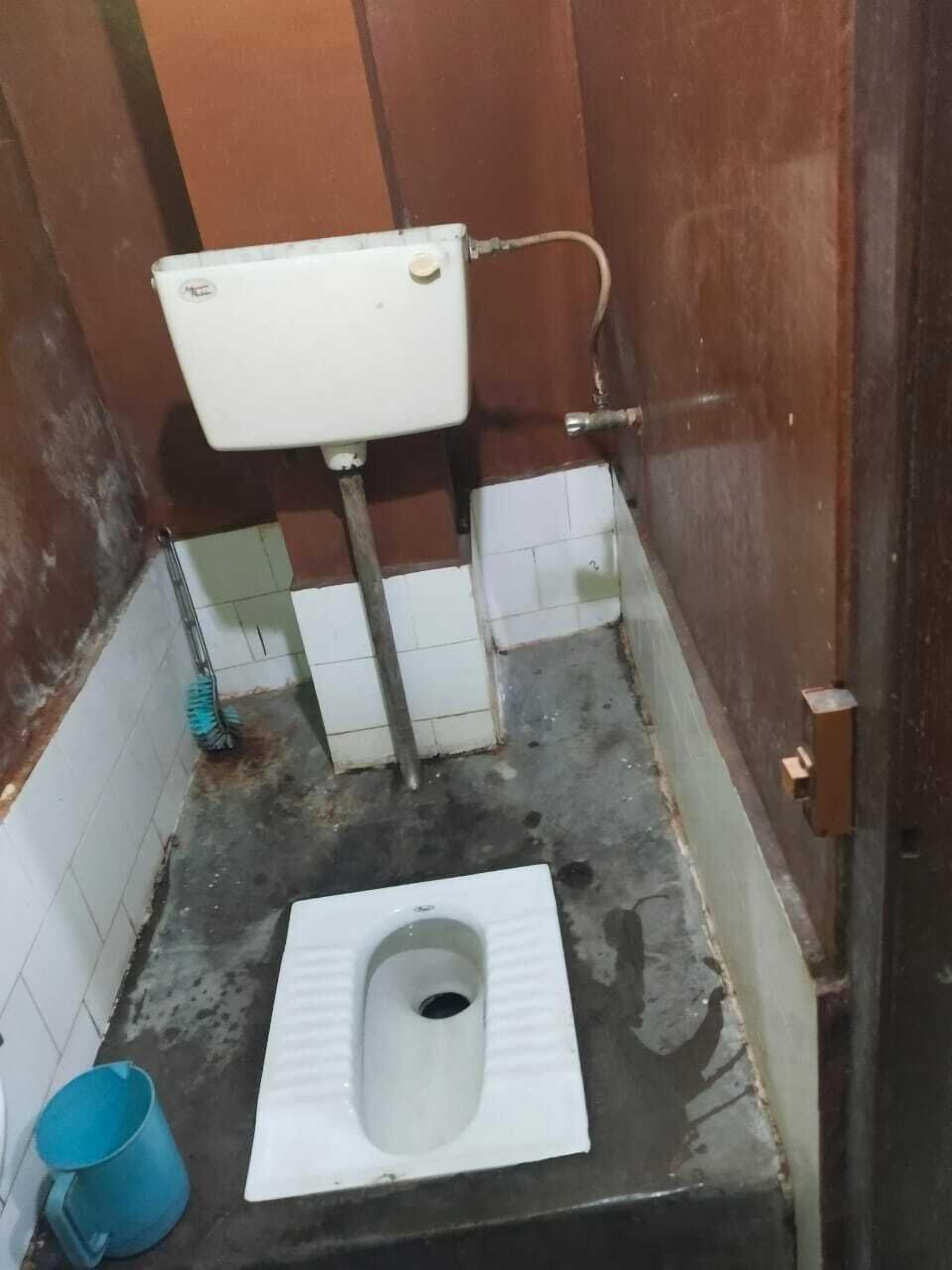Un baño pequeño y sencillo con un inodoro a piso, un cubo azul y un tanque de inodoro blanco.