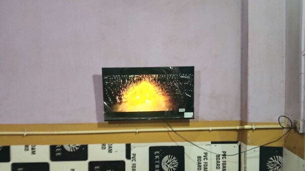 Un téléviseur fixé au mur diffuse des images d’un incendie. En dessous du téléviseur, on peut voir des boîtes étiquetées « PANNEAUX EN MOUSSE DE PVC ».