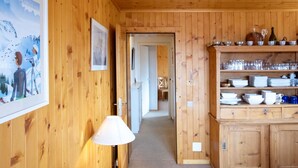 A wooden interior with a hallway leading to a kitchen area. There are framed pictures on the walls, a lamp, and a cabinet with dishes and glasses. - Beautiful apartment for 2 guests with WIFI and balcony (Ollon)