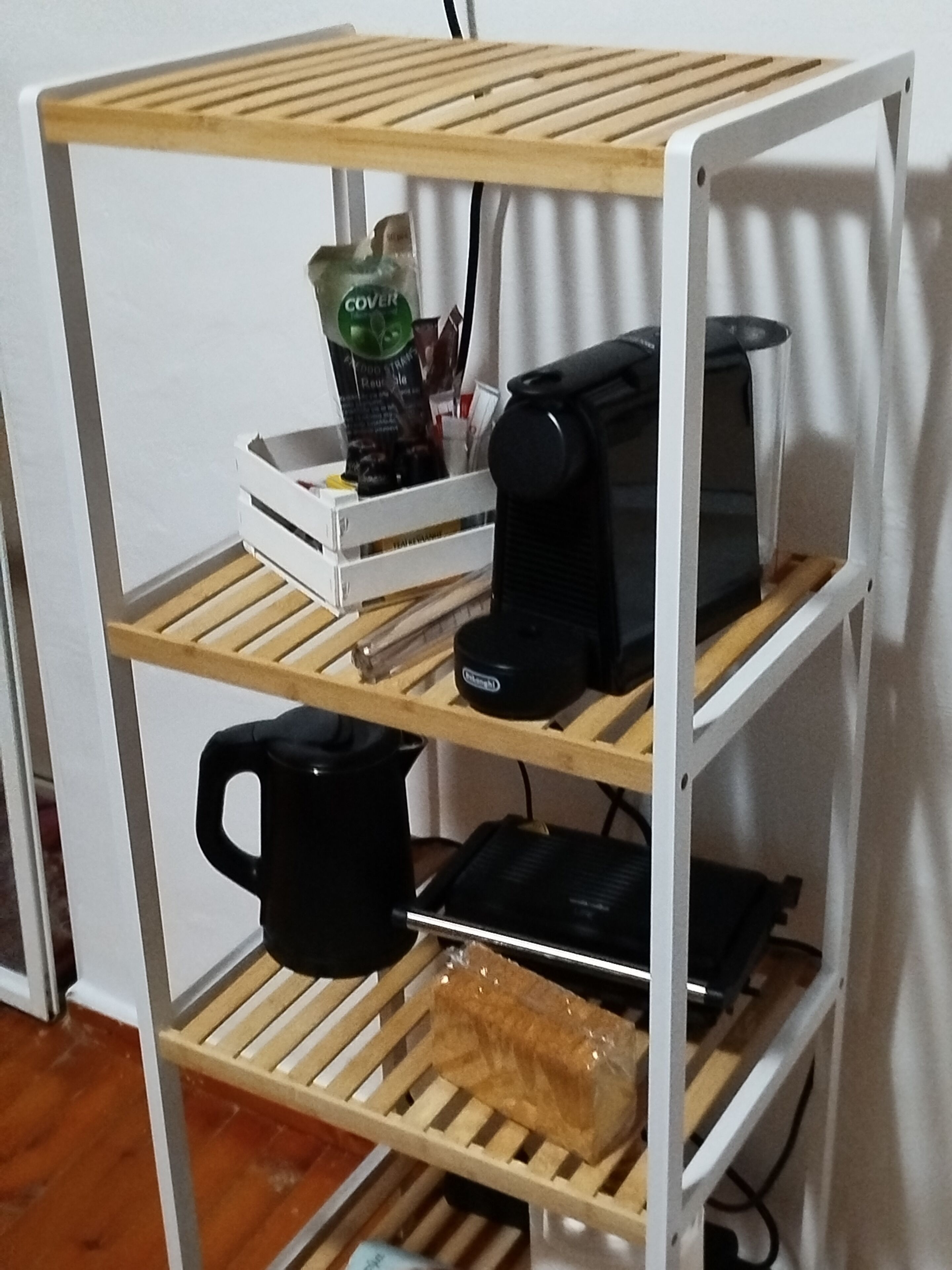 Ein weißes, metallenes Regal mit drei Holzböden. Der oberste Boden beherbergt eine Kaffeemaschine und eine Kaffeetüte. Der mittlere Boden zeigt einen schwarzen Wasserkocher und eine weiße Kiste. Der unterste Boden enthält eine schwarze Tasse und einen in Plastik verpackten Artikel.