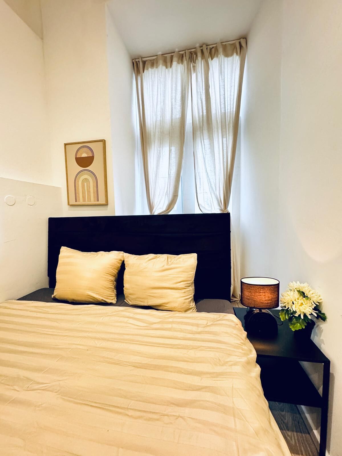 Un dormitorio con cama, dos almohadas, una mesita de noche con lámpara, un florero con flores y un cuadro colgado en la pared.