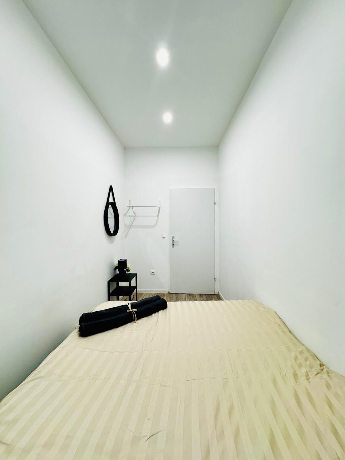 Ein kleiner, einfach eingerichteter Raum mit Holzboden, einem Bett, einer Tür und einer schwarzen Tasche an der Wand.