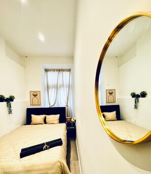 Ein modernes Schlafzimmer mit einem großen Spiegel, einem Bett mit beige farbenem Bettzeug, einem Nachttisch mit Lampe sowie zwei an der Wand befestigten Bilderrahmen.