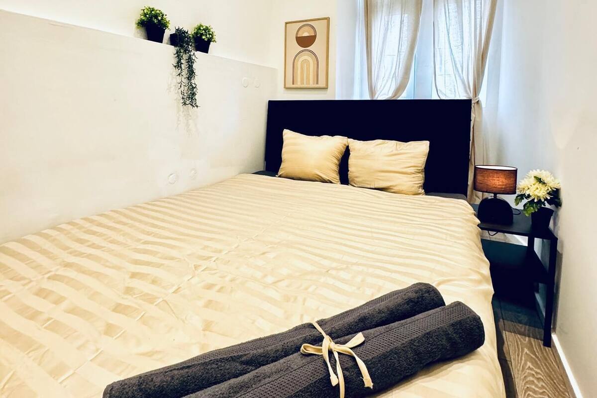 Uma cama bem arrumada com uma toalha enrolada, uma cabeceira escura, uma pequena mesa de cabeceira com abajur e uma planta fixada na parede.