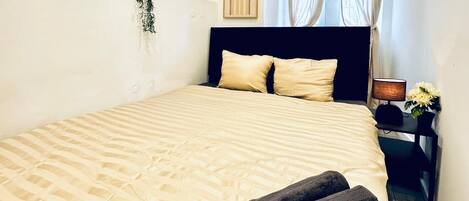A neatly made bed with a rolled towel, a framed artwork on the wall, and a small table with a lamp and flowers.