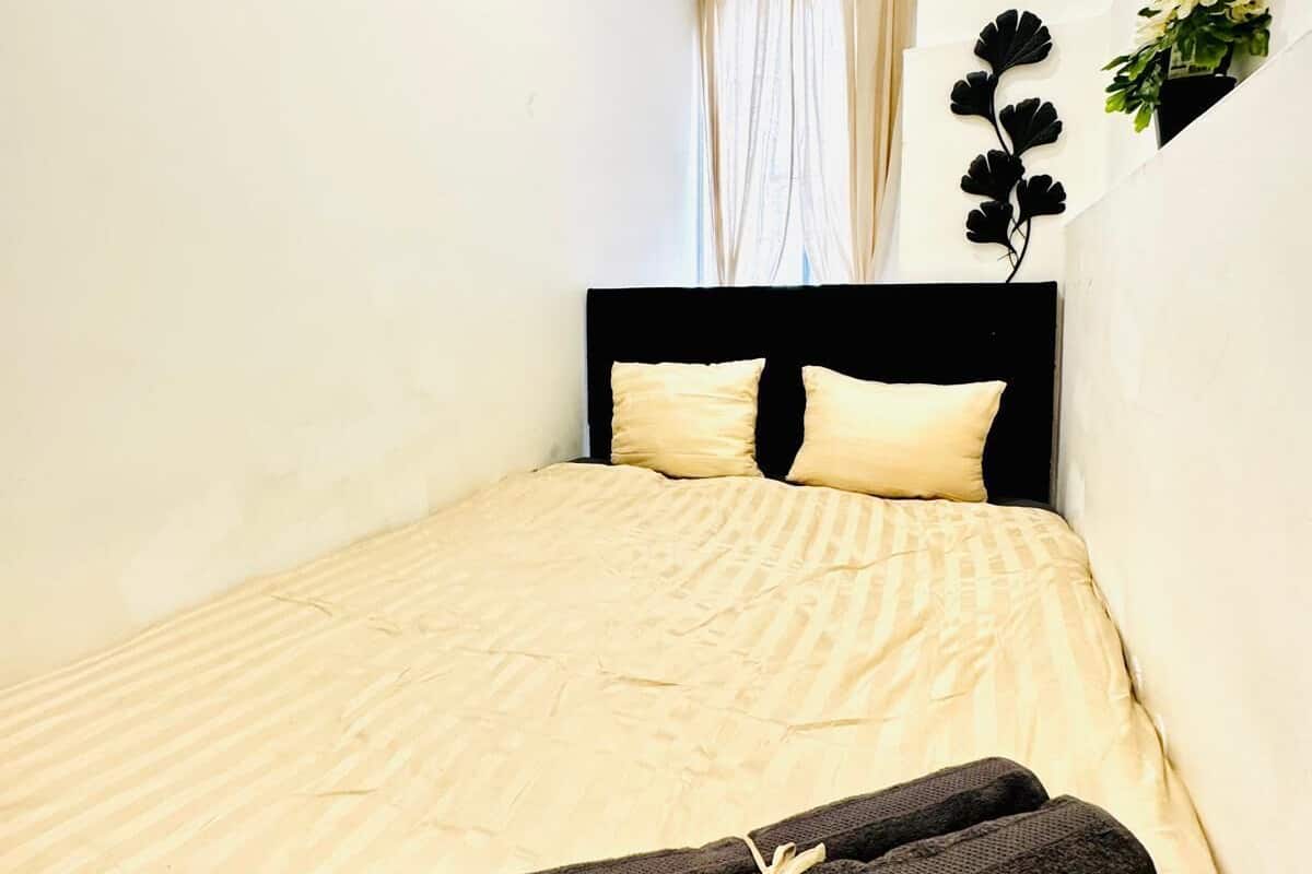 Uma cama pequena e bem arrumada, com um cobre-leito bege e duas almofadas, uma cabeceira preta e um par de toalhas pretas sobre a cama.