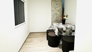 Une pièce étroite avec un plancher en bois, une fenêtre ornée de rideaux sombres, une table recouverte d’une nappe à motifs et deux tabourets noirs.
