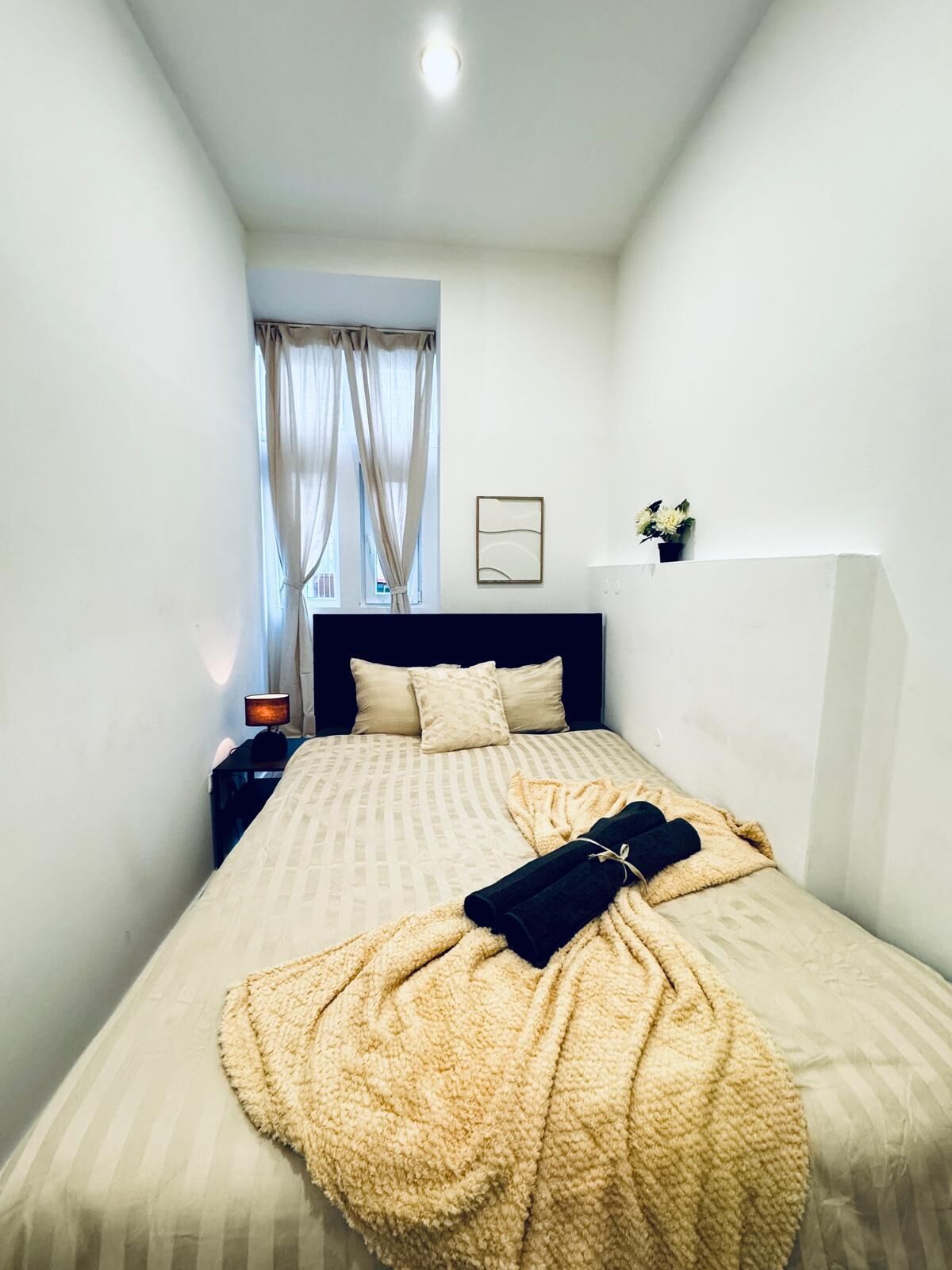 A narrow hotel room with a bed, a nightstand, a lamp, and a window with curtains.