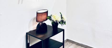 Una habitación con una estantería metálica negra que contiene una lámpara y una planta pequeña. Se ve una pared blanca con un espejo y detalles decorativos.