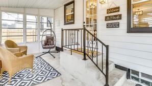 A porch with wicker chairs, a swing chair, a patterned rug, and a staircase with a metal railing.