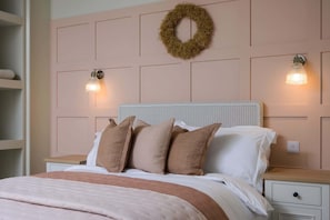 A bedroom with a bed, two bedside tables, a nightstand, a wall-mounted light fixture, and a decorative wreath.