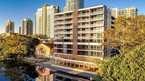 A modern multi-story building with balconies, situated near a body of water with trees. - Prime Loc! 1BR Apt with Parking Heart of Surfers (Surfers Paradise)