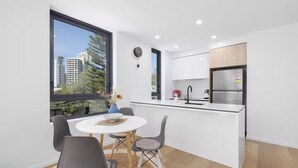 A modern kitchen with white cabinets, a black faucet, and a stainless steel refrigerator. There is a dining area with a round table and two chairs, and a large window offering a view of buildings and trees. - Prime Loc! 1BR Apt with Parking Heart of Surfers (Surfers Paradise)