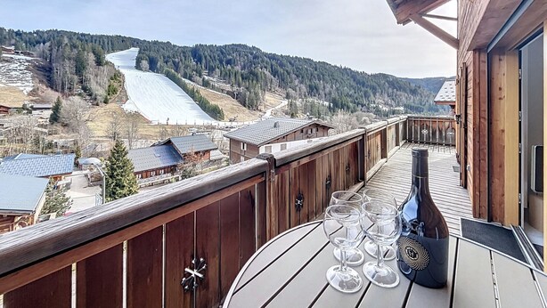 Un balcón de madera con una mesa, copas de vino y una botella, con vistas a una pista de esquí y montañas boscosas.