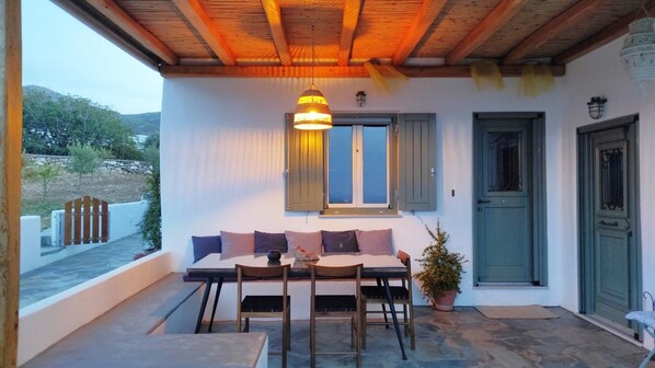 A cozy outdoor seating area with a table and chairs, a hanging light fixture, and a door with a green shutter.
