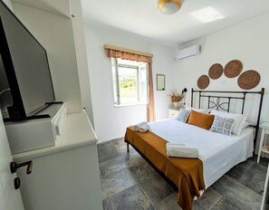 A bedroom with a bed, a bedside table, a window with curtains, a wall-mounted air conditioner, and a view of the outdoors.