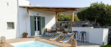 Un espace piscine doté de chaises longues en bois, d’un mur en pierre et d’un coin salon couvert avec une table et des chaises.