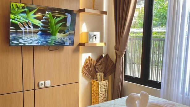 A hotel room with a bed, a flat-screen TV mounted on the wall, a window with curtains, and a small shelf with a decorative item. - DIRUMA SADJA HOME VILLA (Ngemplak)