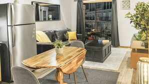 Una sala moderna con una mesa de comedor de madera, sillas tapizadas en gris, un sofá de cuero negro con cojines amarillos, un refrigerador de acero inoxidable y vistas a un balcón con mobiliario exterior.