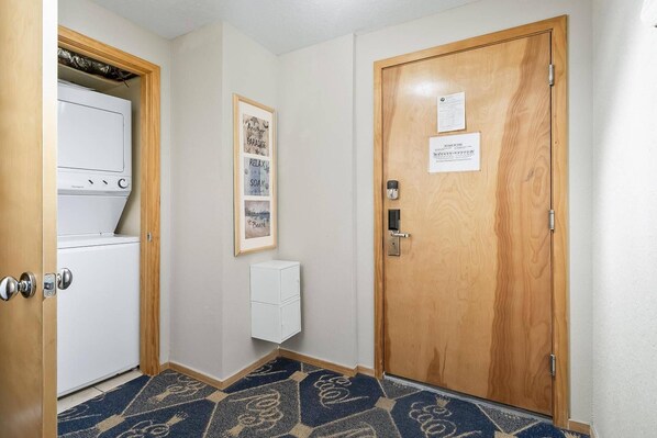 Un pasillo con una puerta de madera, una pared blanca con un cartel enmarcado y un electrodoméstico blanco con panel de control.