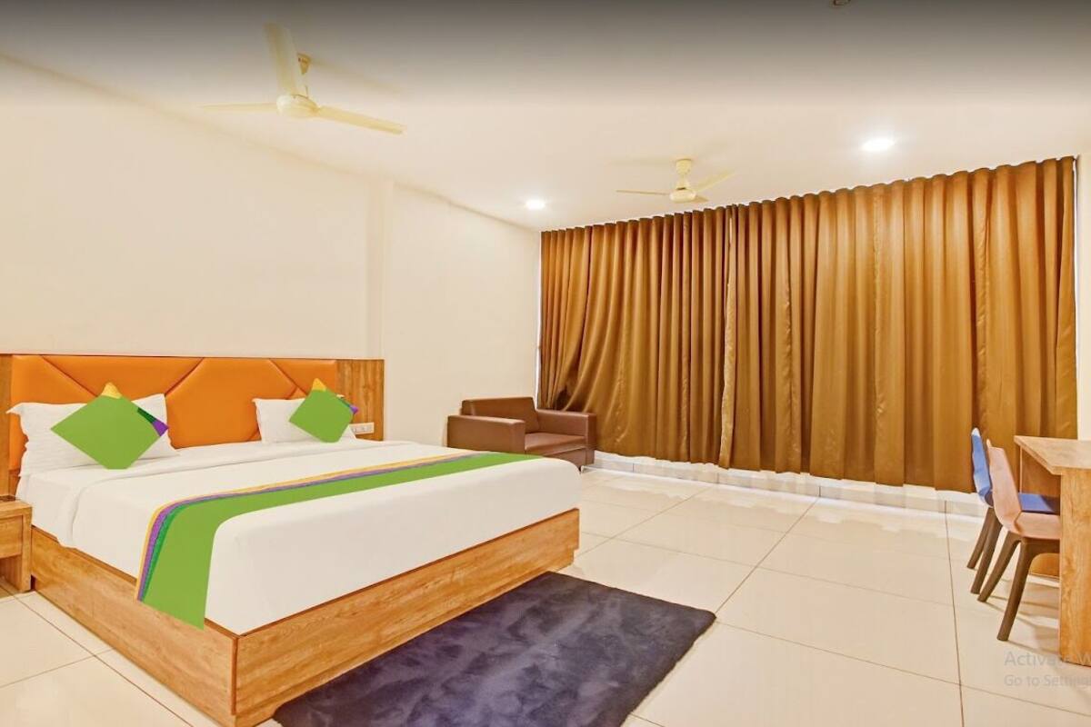 Quarto de hotel com uma cama grande, uma escrivaninha, uma cadeira, uma mesinha de cabeceira e uma janela com cortinas.