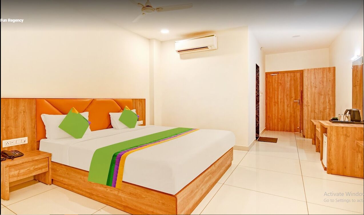A hotel room with a wooden bed, white walls, an air conditioning unit, a wooden wardrobe, and a small desk with a chair.