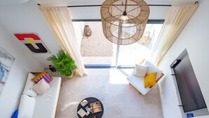 Een moderne woonkamer met een bank, een salontafel en een televisie. Er is een groot raam met gordijnen, een hanglamp en een potplant.