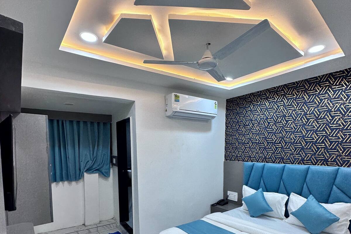 A modern hotel room with a geometric ceiling design, a bed with blue and white bedding, a bedside table with a tray, and a patterned wall.