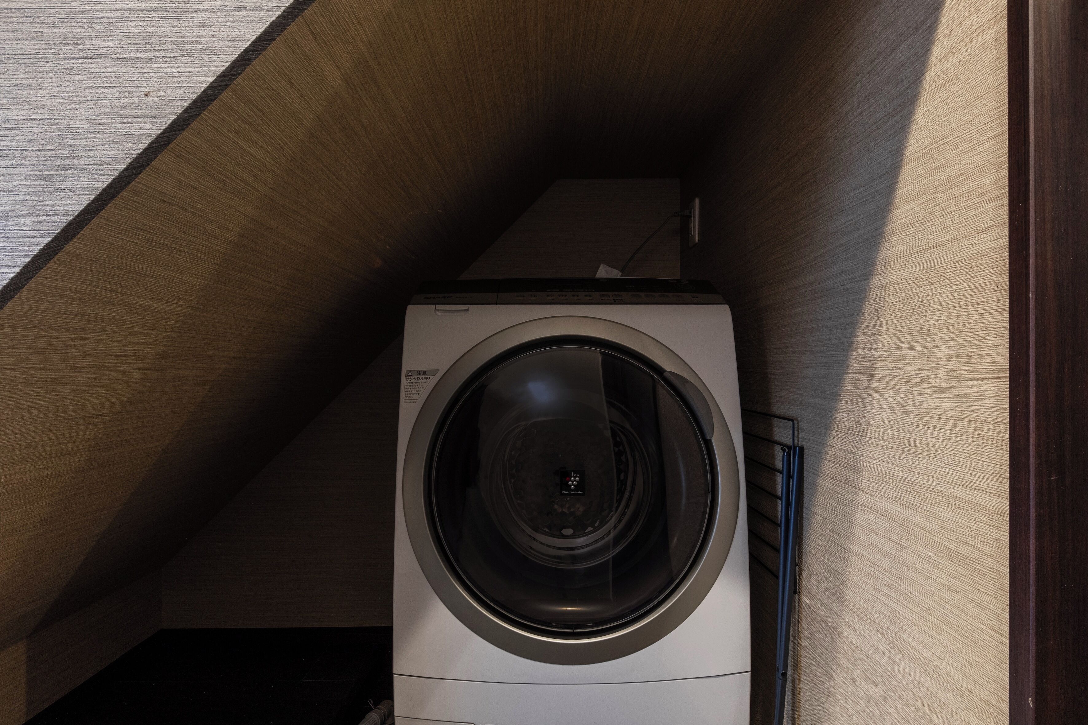 A front-loading washing machine in a corner with a wooden ceiling and a visible power outlet.
