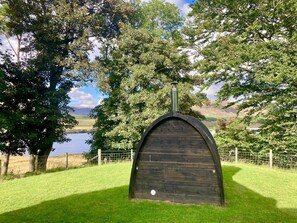 Estructura de madera con chimenea, rodeada de árboles y un área cubierta de césped, con vista a un lago y colinas al fondo.