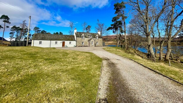A stone house with a gravel driveway leading to a white building with a red door. - Sherrabeg Cottage -  a cottage that sleeps 8 guests in 4 bedrooms (Laggan)