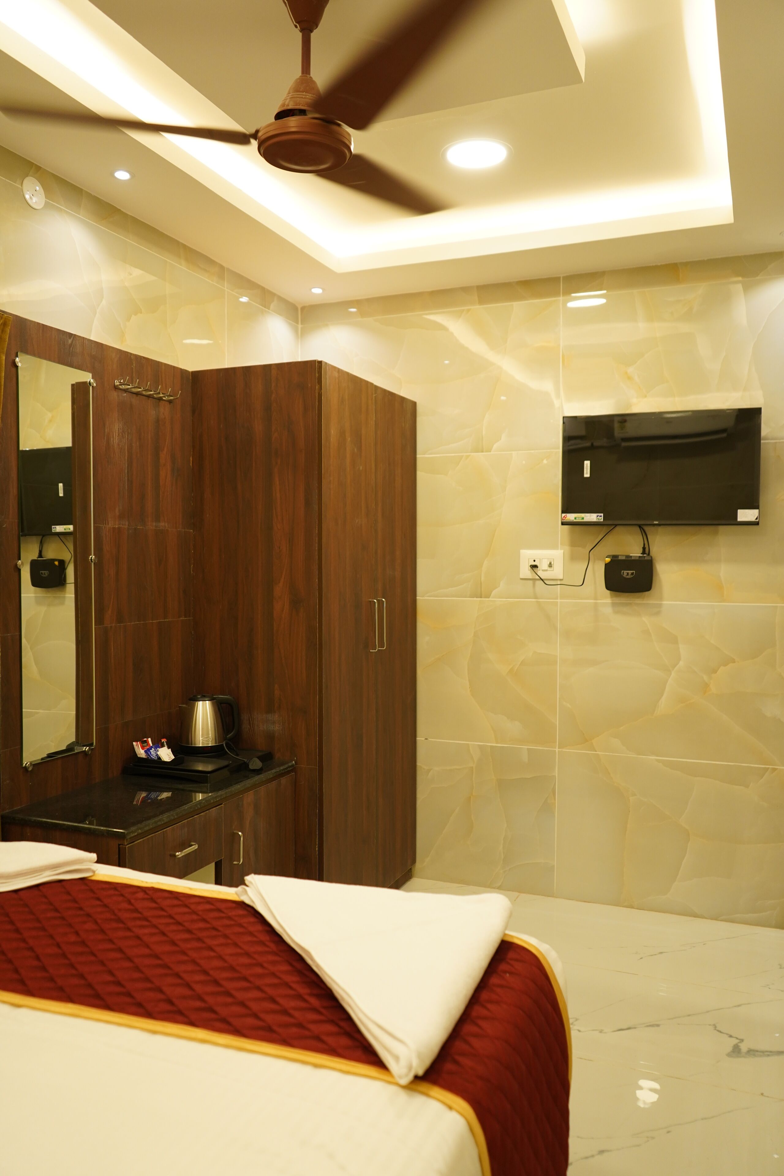 A hotel room with a bed, a wardrobe, a mirror, a ceiling fan, a TV mounted on the wall, and a coffee maker on the nightstand.